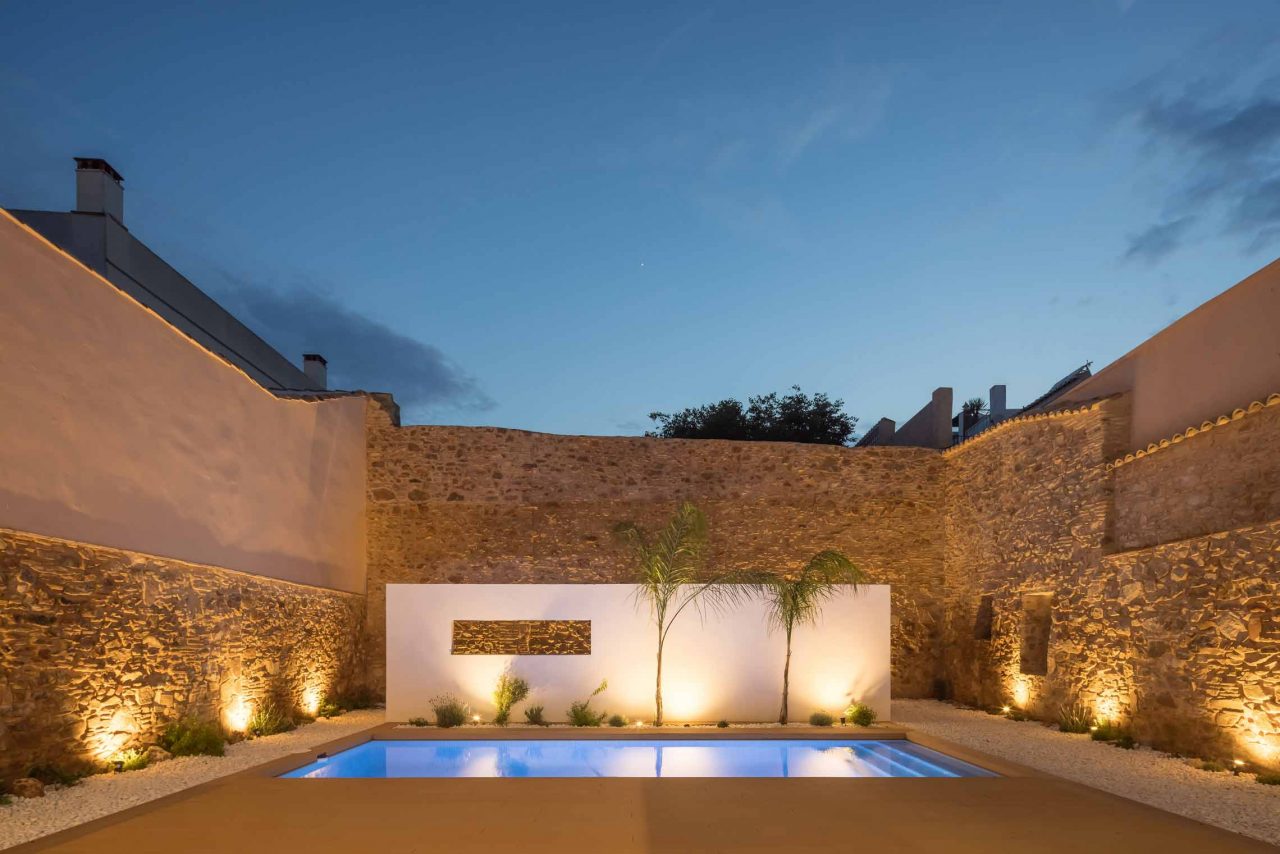 Rehabilitación y reforma Casa A_M Puzol Valencia - Vista nocturna del patio interior con piscina y muro separando zona de barbacoa rodeado por muros de mamposteria originales